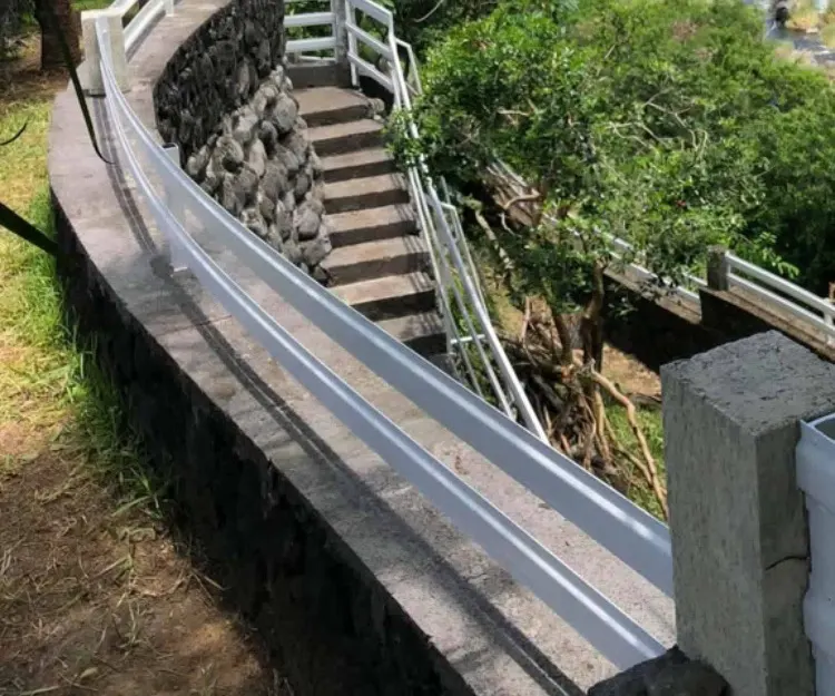 Un escalier aux rampes blanches qui descend à flanc de falaise, entre le mur de pierre et la verdure